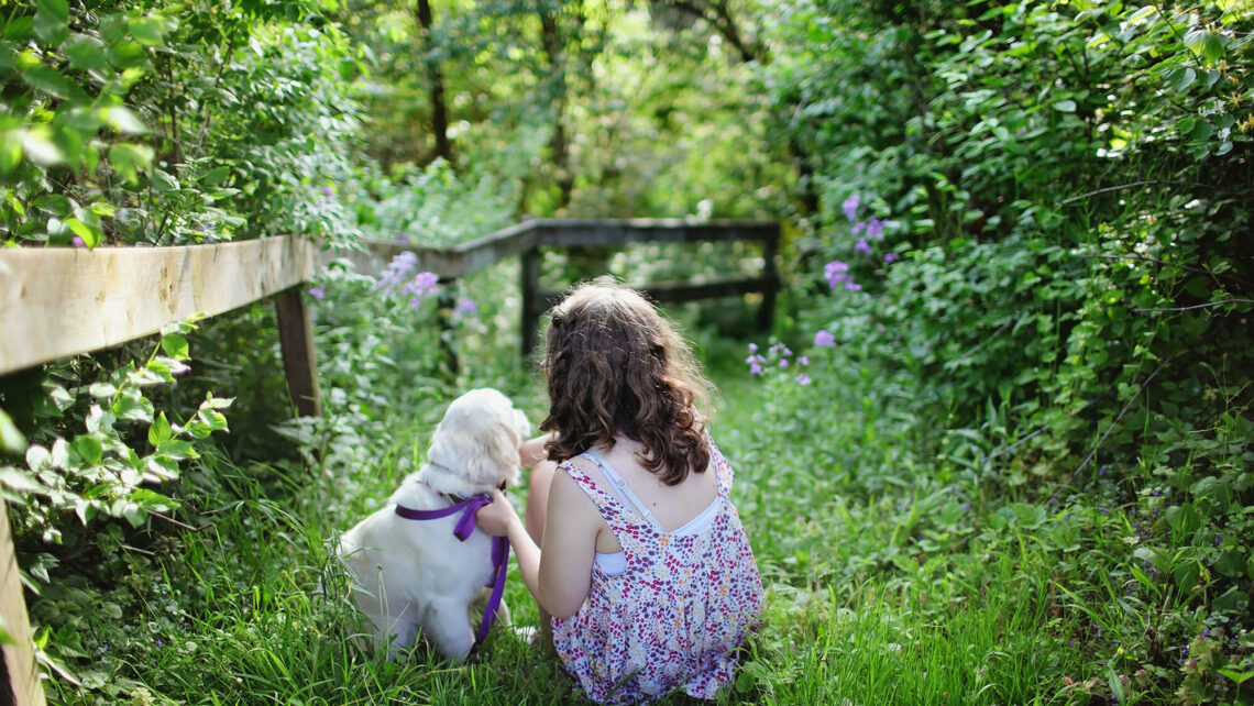 A young girl in a purple dress holds the purple colour of a golden retriever puppy as they look at a lush green garden during the Spring.